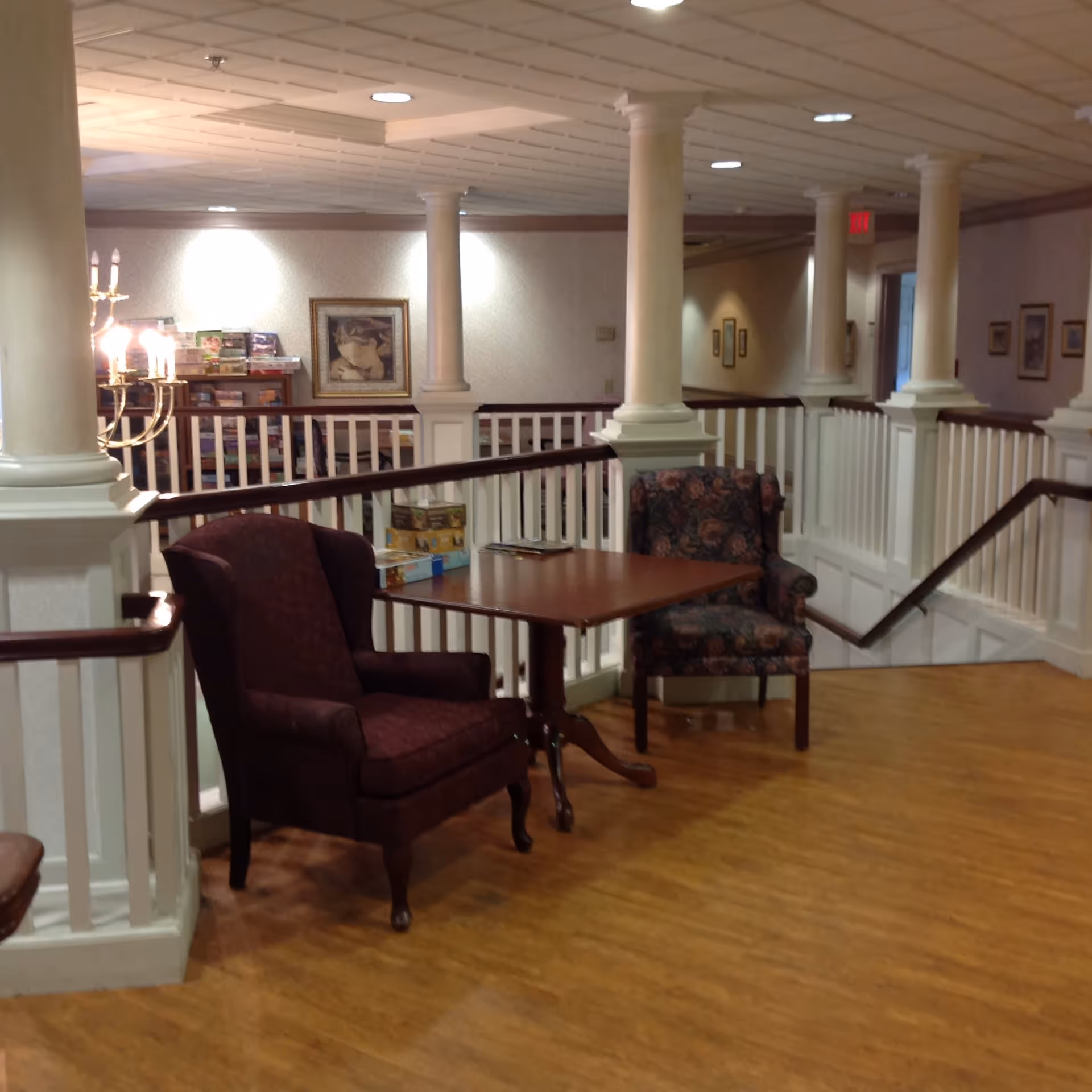 An interior common area with two upholstered armchairs and a wooden table between them, situated near white railing and columns. The space has wooden flooring, framed artwork on the walls, and soft lighting.