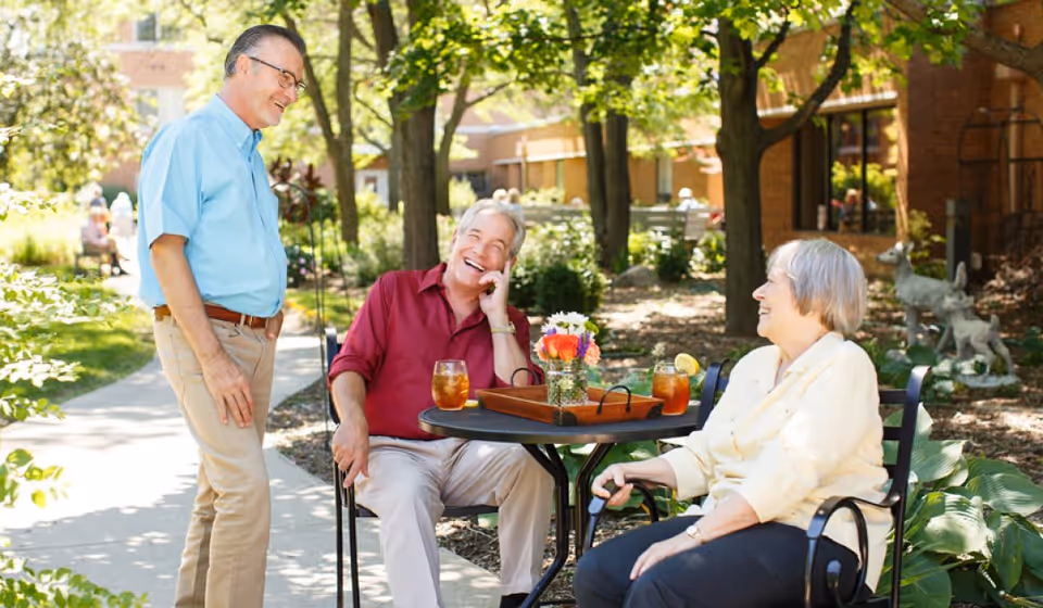 Three older adults laugh and talk around a small outdoor patio table in a tree-shaded courtyard of a senior living community.