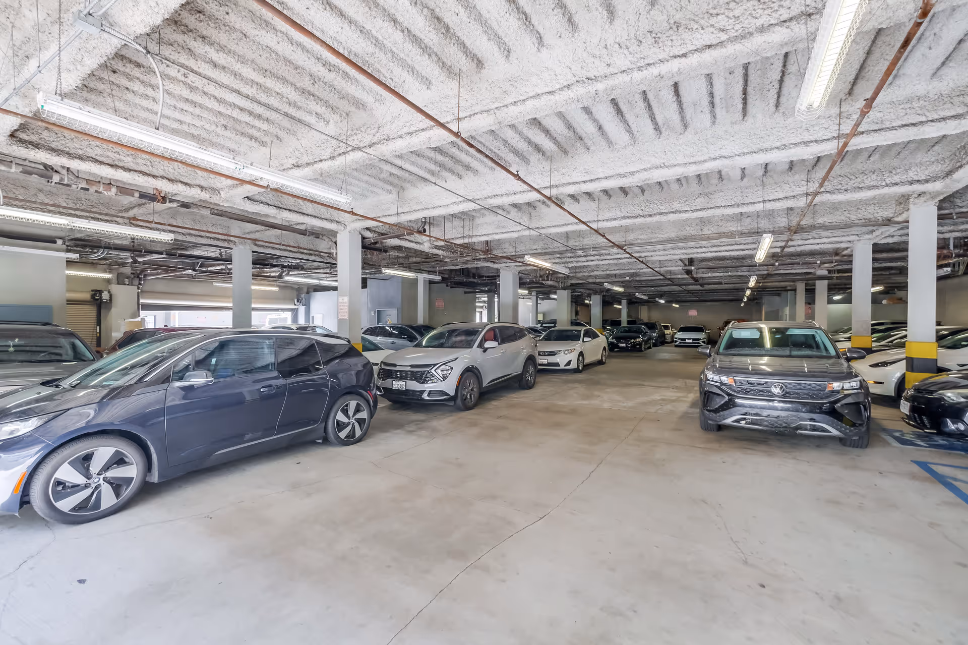 Indoor parking garage with multiple parked cars including a black Volkswagen and other various vehicles under a ceiling with exposed pipes and fluorescent lighting.