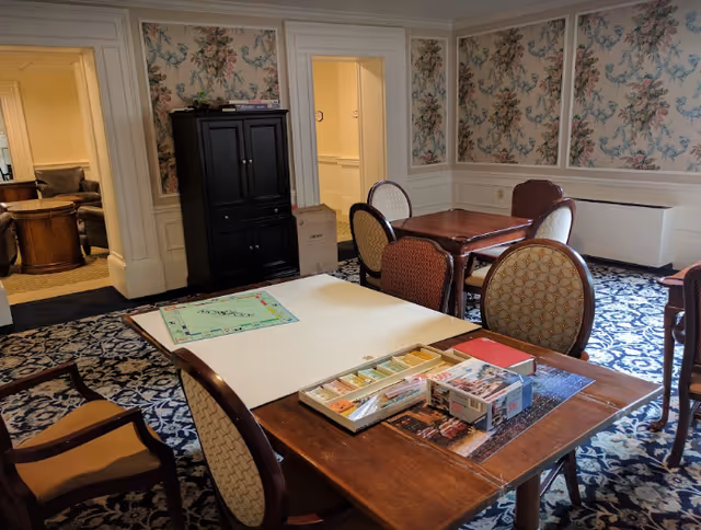 A cozy interior room with floral wallpaper and patterned carpet, featuring several wooden tables and chairs. One table has a Monopoly board game set up with game pieces and cards. In the background, there is a black cabinet and an open doorway leading to another room with additional seating.