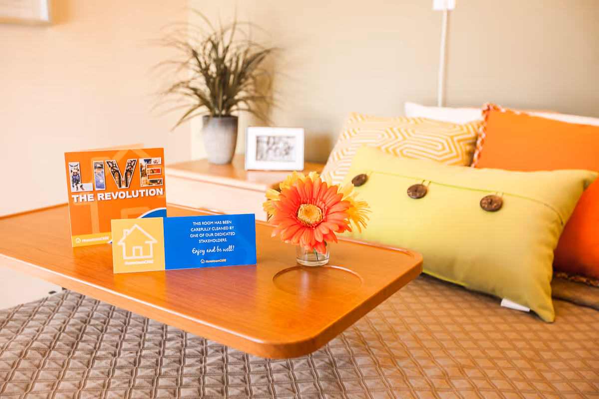 A wooden overbed table with colorful cards and a small vase of orange flowers placed on a bed with decorative pillows.