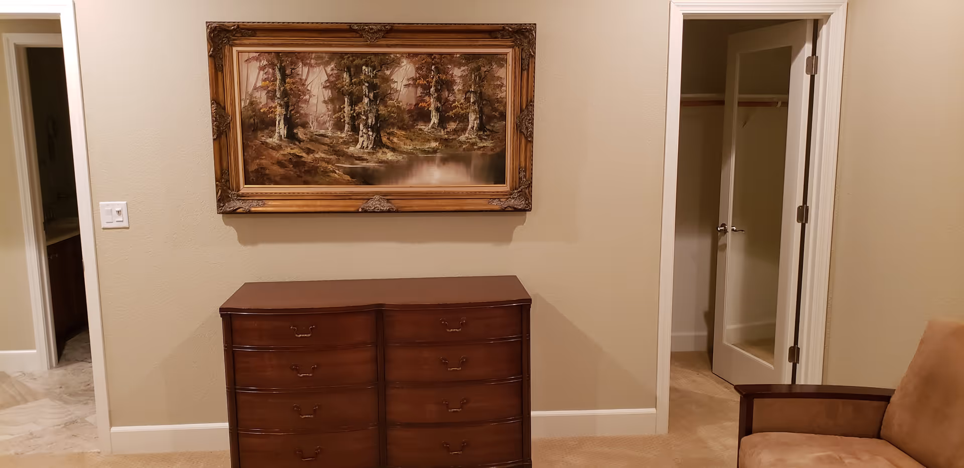 Wooden dresser against a beige wall with a framed landscape painting above, doorways and a closet on either side, and an armchair at right.