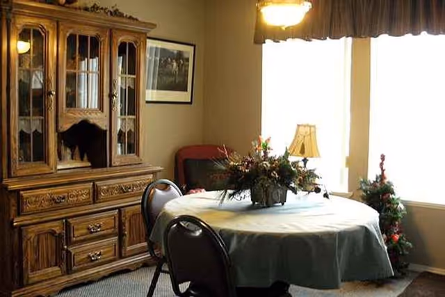 A dining room with a round table covered with a light-colored tablecloth and a floral centerpiece. Four chairs surround the table. To the left, there is a wooden china cabinet with glass doors and drawers. A framed picture hangs on the wall above a red cushioned chair. On the right side, there is a window with curtains, a table lamp, and a small decorated Christmas tree.