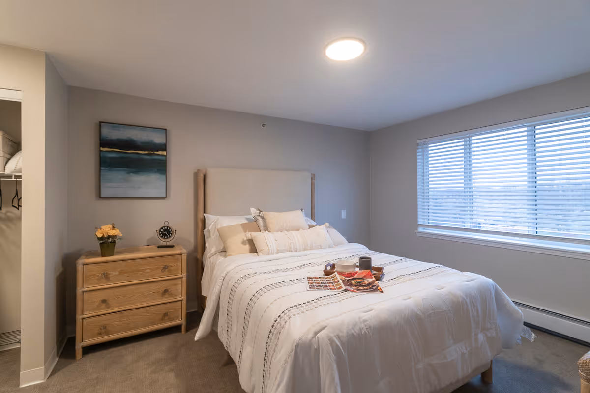 A cozy bedroom with a neatly made bed featuring white and beige pillows and a white comforter with subtle patterns. A wooden nightstand with three drawers sits next to the bed, holding a small clock and a potted plant with yellow flowers. A framed abstract painting hangs on the wall above the nightstand. A large window with horizontal blinds lets in natural light, and an open closet is partially visible on the left side of the image.