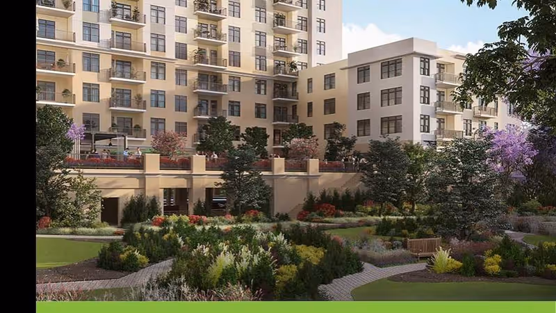 A landscaped garden area with various shrubs, trees, and flowers in front of a multi-story residential building with balconies and large windows under a partly cloudy sky.