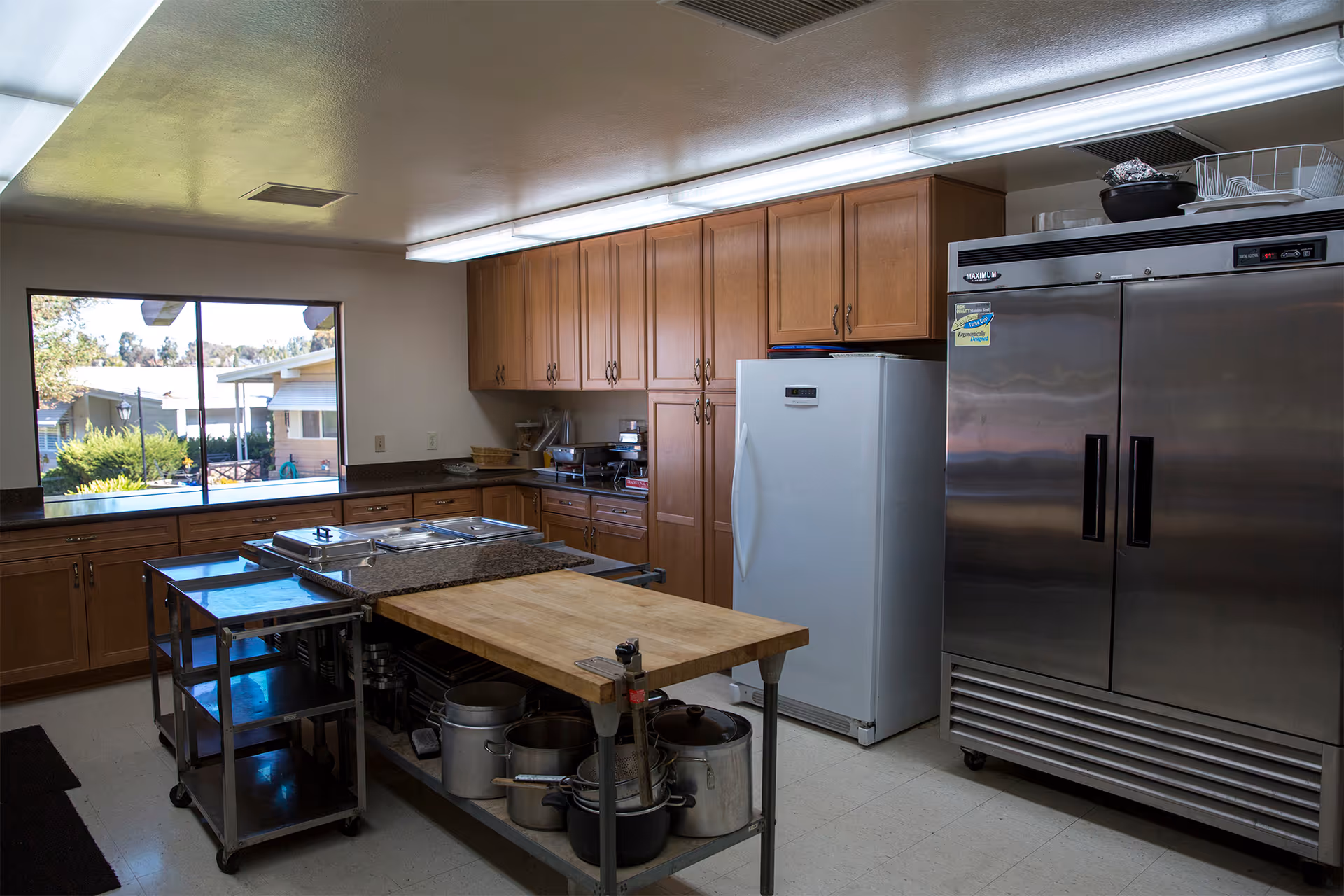A commercial-style kitchen with wooden cabinets, a large stainless steel refrigerator, a white freezer, a metal rolling cart, and a central island with a wooden cutting board and various pots and pans underneath. A window shows an outdoor view of neighboring buildings and greenery.