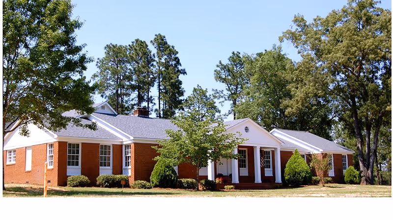 A single-story brick building with white trim and a gray roof, surrounded by trees and shrubs under a clear blue sky.