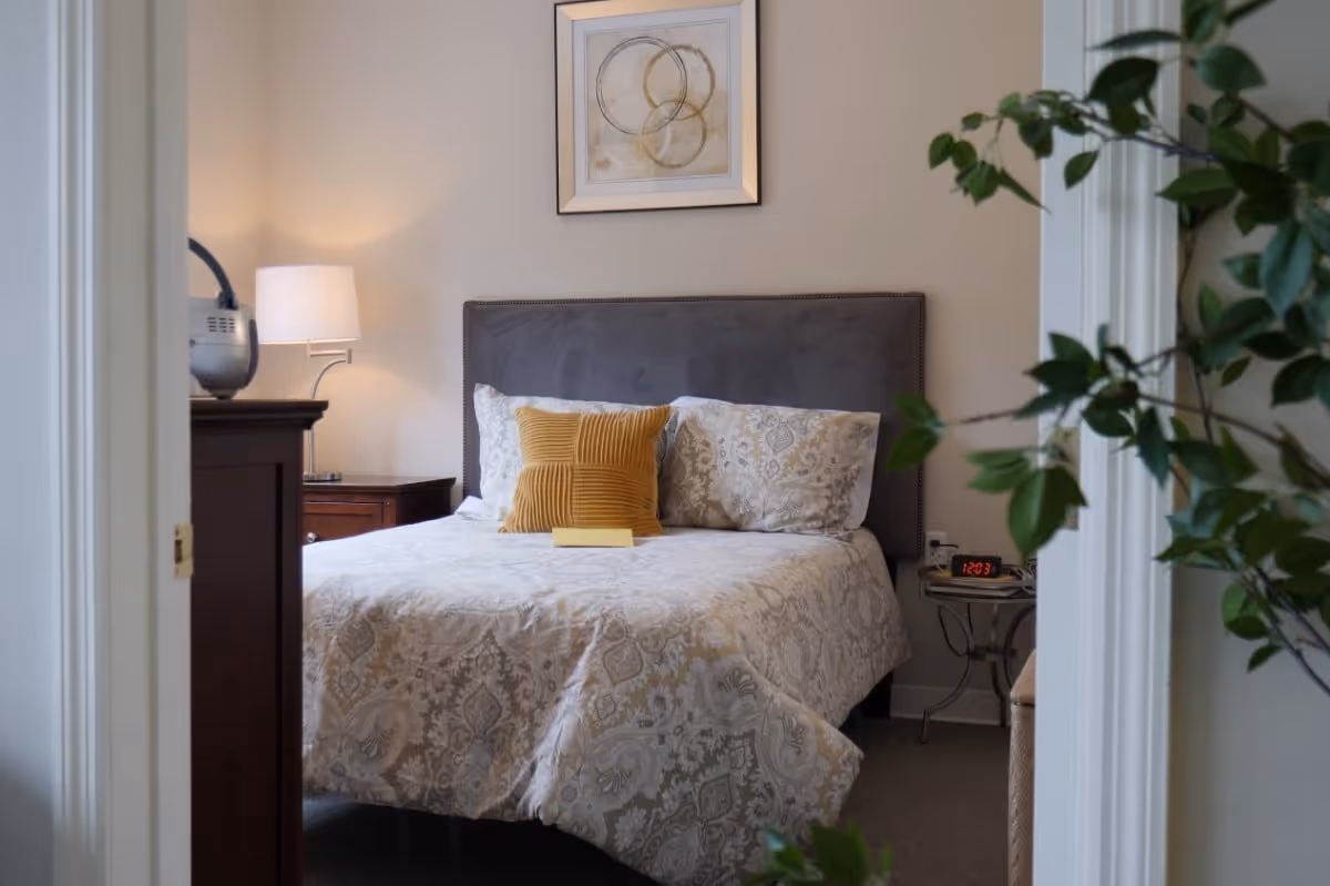 View through a doorway into a bedroom with a neatly made bed featuring patterned bedding and two pillows, one mustard yellow and one patterned. A bedside table with a lamp and an alarm clock showing 12:03 is visible. A framed abstract artwork hangs above the bed, and a leafy green plant is partially visible in the foreground.