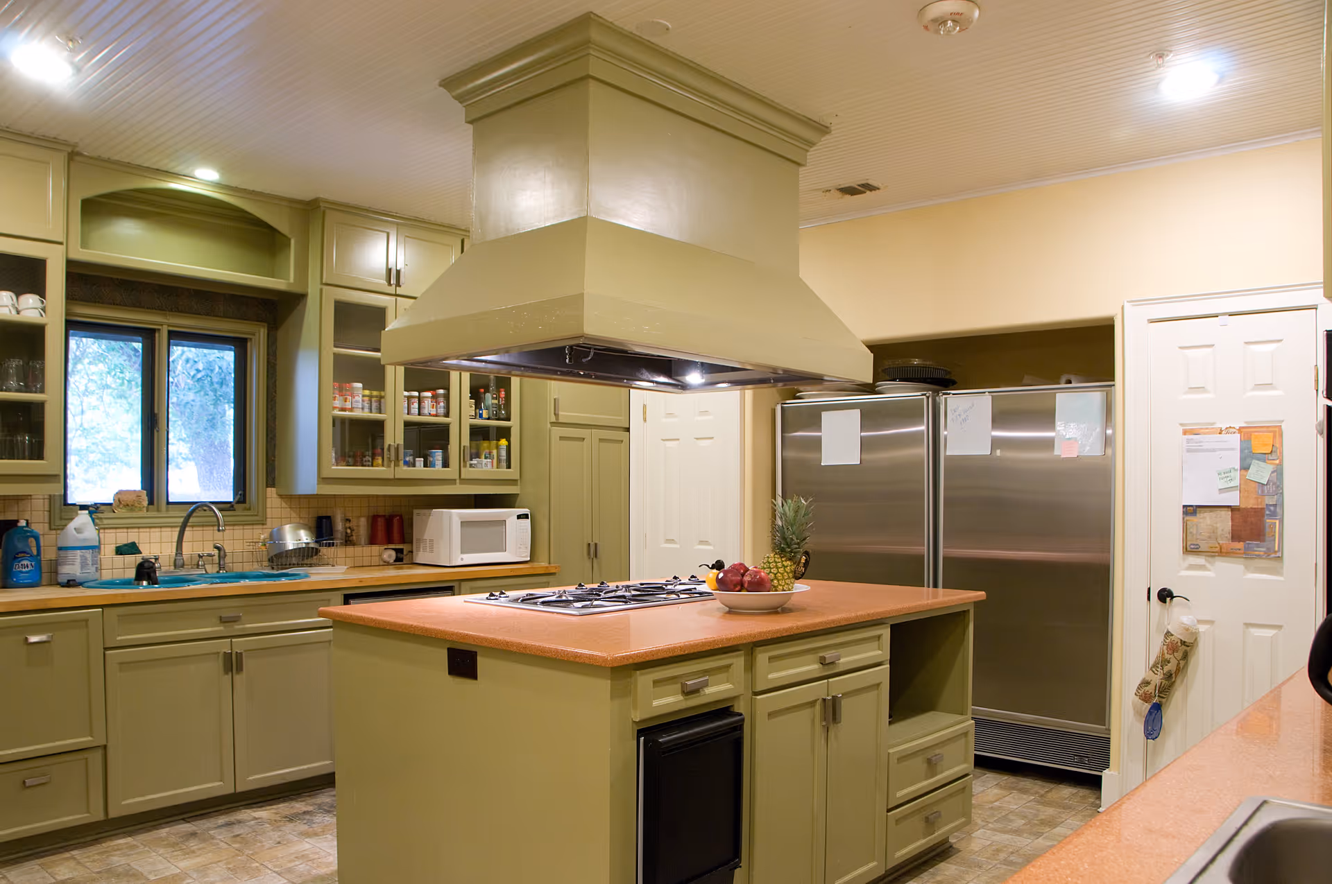 A spacious kitchen with green cabinetry and a central island featuring a built-in stovetop and a large overhead vent hood. The kitchen includes a double stainless steel refrigerator, a microwave, a sink under a window, and various kitchen items such as a bowl of fruit on the island and cleaning supplies near the sink.