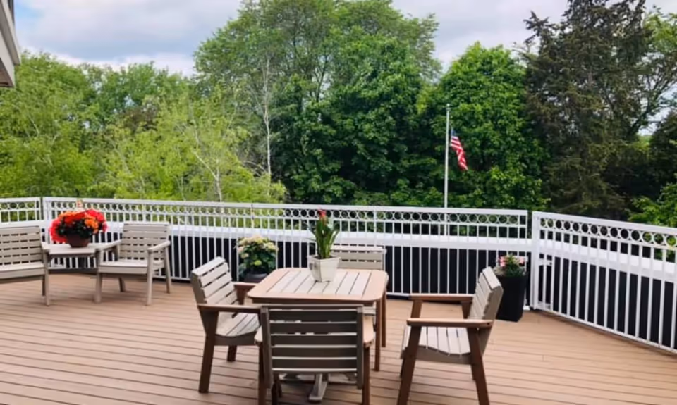 Outdoor patio area with wooden deck flooring, featuring a square table with four chairs around it and a smaller table with two chairs. Several potted plants with colorful flowers are placed around the patio. A white railing surrounds the patio, and lush green trees are visible in the background under a partly cloudy sky. An American flag is also visible on a flagpole beyond the railing.