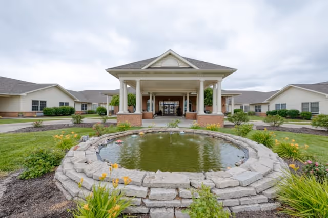 Front exterior view of Cedarhurst Senior Living of Granite City showing a covered entrance with columns, a circular stone-bordered pond with water and surrounding landscaping including green grass and flowering plants under a cloudy sky.