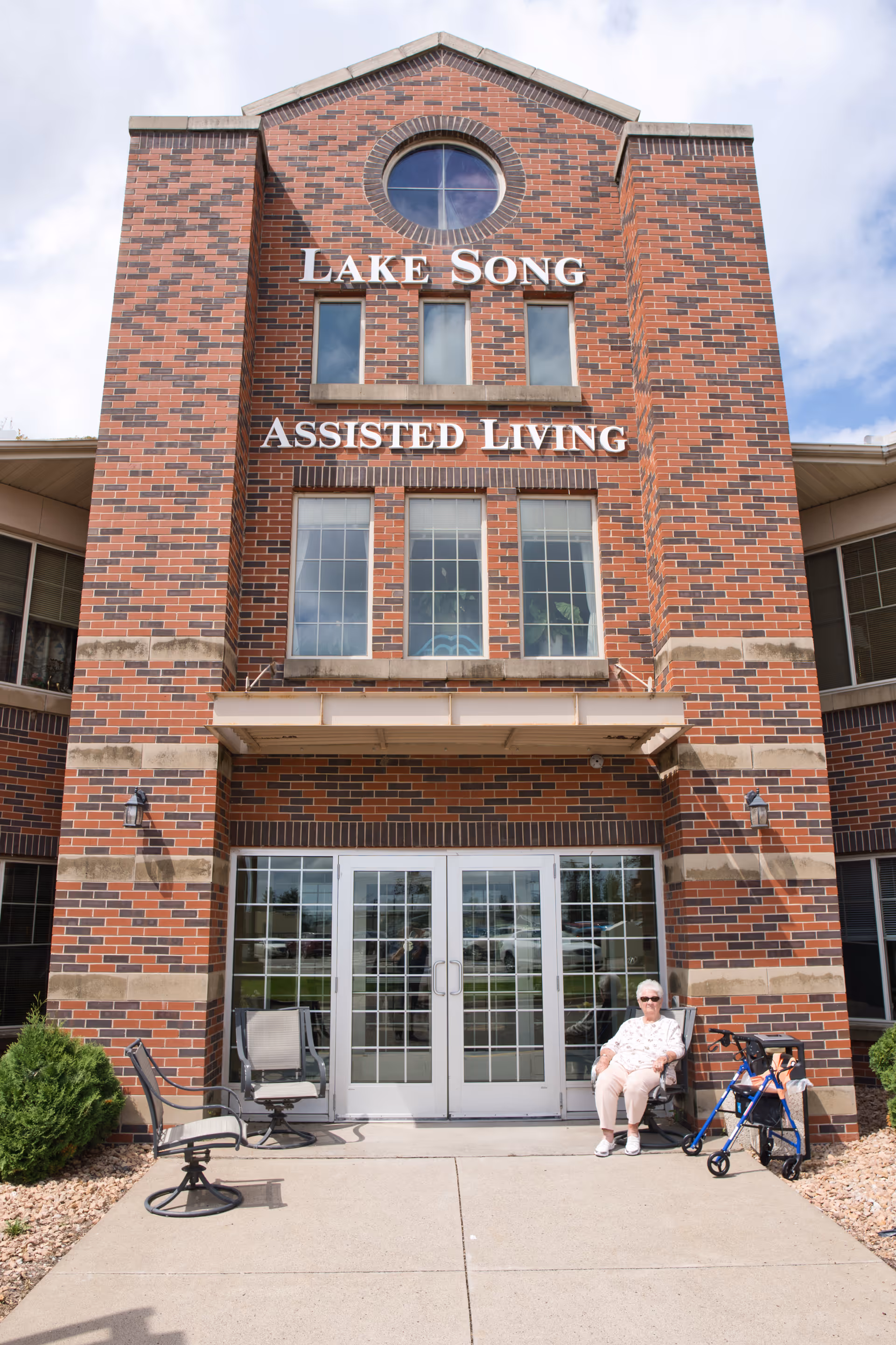 Front entrance of the brick Lake Song Assisted Living building with double glass doors and an older adult seated beside a walker.