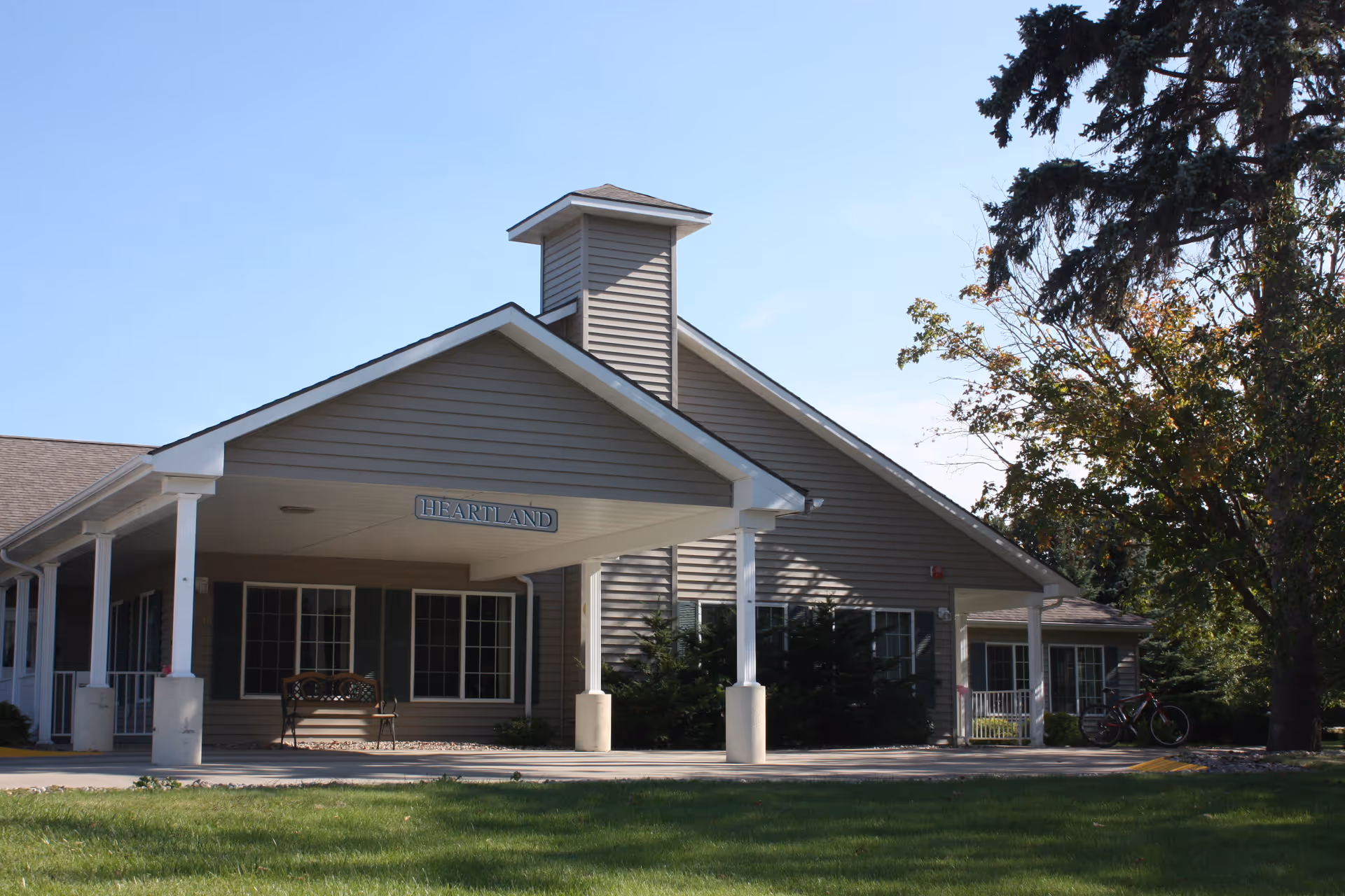 Front entrance of a single-story senior living building with a covered porch, columns, bench, and a 'HEARTLAND' sign, with lawn and trees in front.
