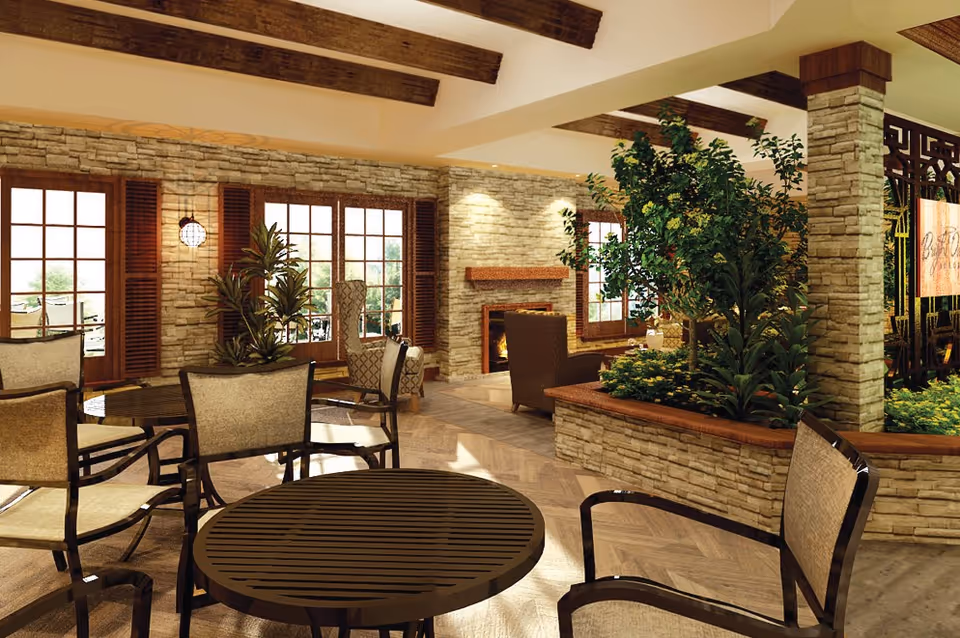 A cozy and well-lit common area with stone walls, wooden ceiling beams, and large windows. The room features several round tables with cushioned chairs, a stone fireplace with a fire burning, and a planter filled with green plants. The space has a warm and inviting atmosphere with natural light streaming in.