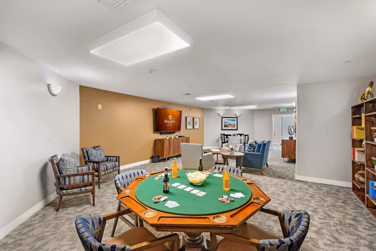 A senior living facility common room with a poker table in the foreground featuring playing cards, poker chips, a bowl of popcorn, and bottles of beverages. The room has comfortable seating including armchairs and sofas, a TV mounted on a tan accent wall displaying the MorningStar Senior Living logo, and a bookshelf with decorative items on the right side.