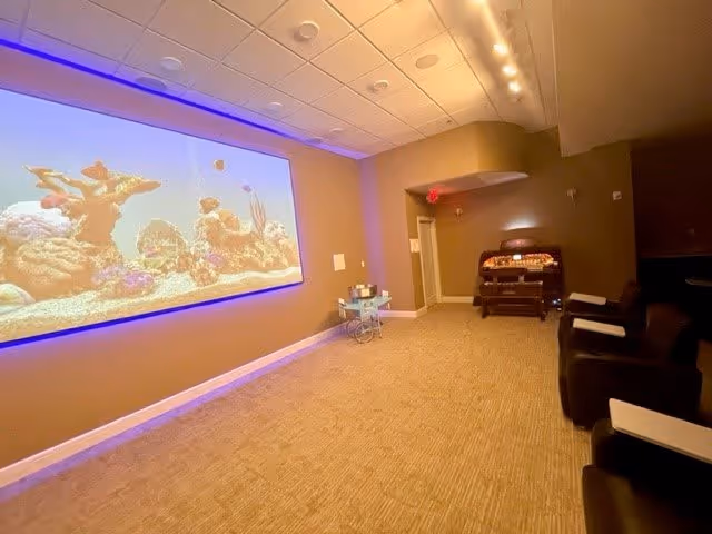 A cozy room with a large screen on the left wall displaying an underwater scene with coral and fish. The room has beige walls and carpeted floor. On the right side, there are several black reclining chairs with white armrests. At the back of the room, there is a small table with items on it and a vintage-style jukebox. The ceiling has recessed lighting and a row of spotlights along the right wall.