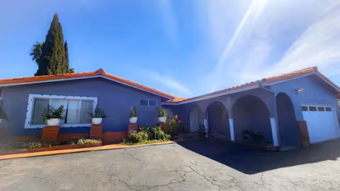 Exterior view of a single-story residential building with a blue-gray facade and red-tiled roof under a clear blue sky. The building features a large window with white trim, several potted plants along the front, an arched covered entrance, and a garage door on the right side. The driveway and surrounding area are paved.