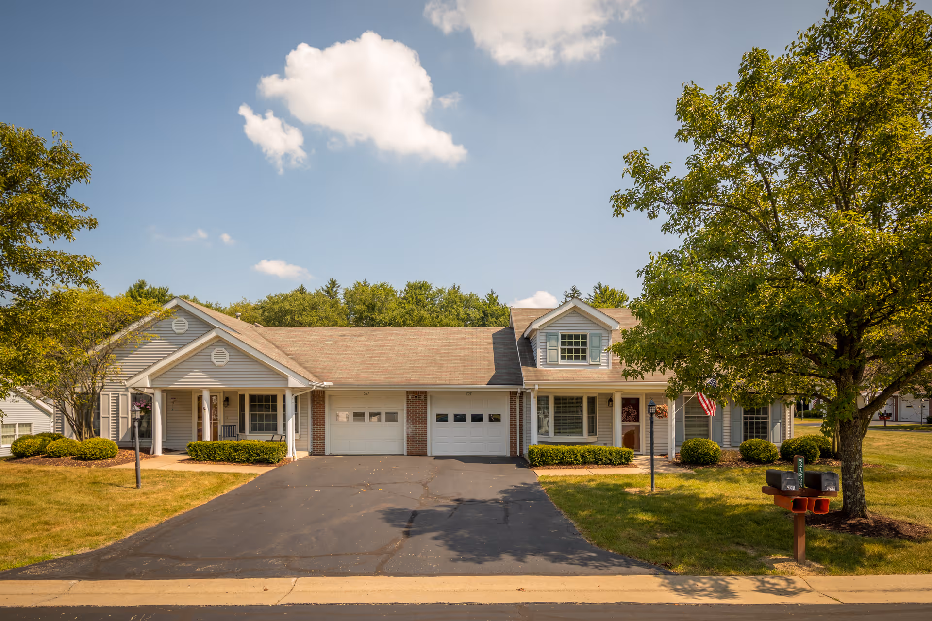 Front exterior of a single-story senior living building with two garage doors, a shared driveway, lawn, trees, and mailboxes.