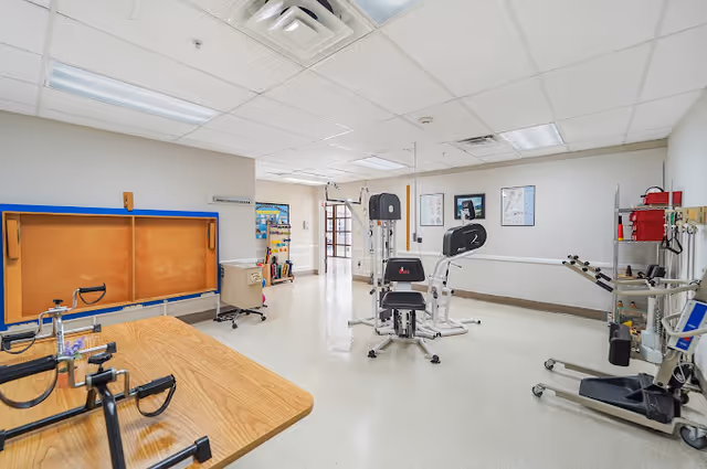 A spacious rehabilitation or physical therapy room with exercise equipment including a multi-station gym machine, parallel bars, and other therapy apparatus. The room has a white ceiling with fluorescent lights, beige walls, and a light-colored floor. There is a bulletin board and some medical charts on the walls.