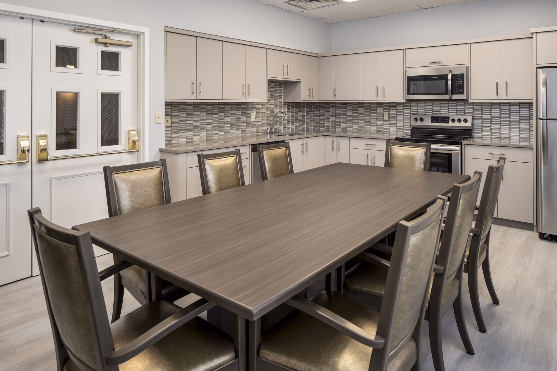 A communal dining area with a long rectangular table and chairs in front of a modern kitchen with cabinets, tiled backsplash, and stainless appliances.