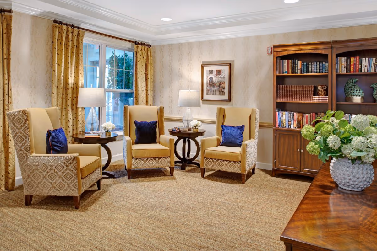 A cozy sitting area with three beige patterned armchairs each with a dark blue cushion, arranged around two small round wooden tables with lamps and decorative items. Behind the chairs is a window with yellow patterned curtains and a framed picture on the wall. To the right is a wooden bookshelf filled with books and decorative pieces. In the foreground, a wooden table holds a white vase with green and white flowers.
