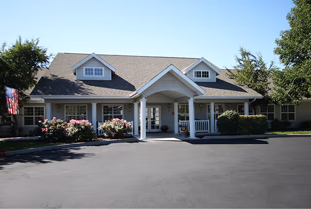 Front exterior view of a single-story senior living facility building with a covered entrance supported by white columns, surrounded by bushes and flowers, under a clear blue sky.