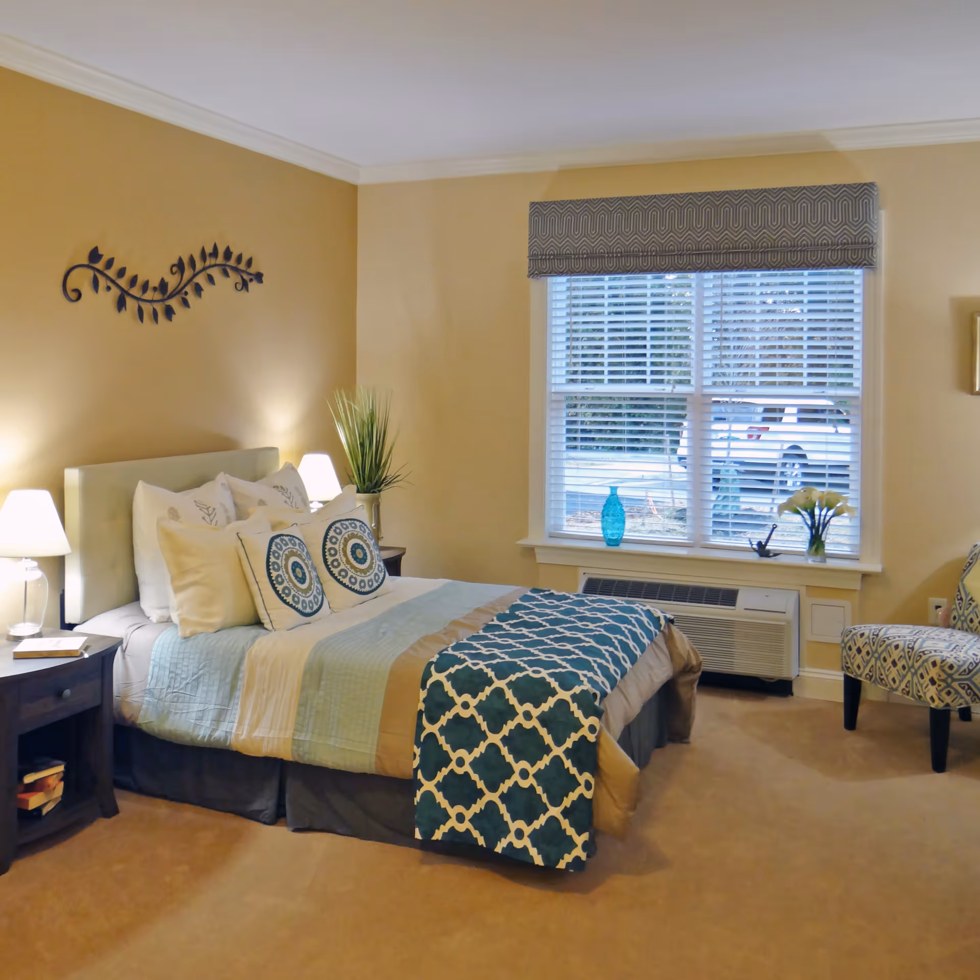 A cozy bedroom with a neatly made bed featuring multiple pillows and a blue patterned throw blanket. There are two bedside tables with lamps, a decorative plant, and a wall decoration above the bed. A window with blinds and a patterned valance lets in natural light, and a patterned armchair is placed near the window.