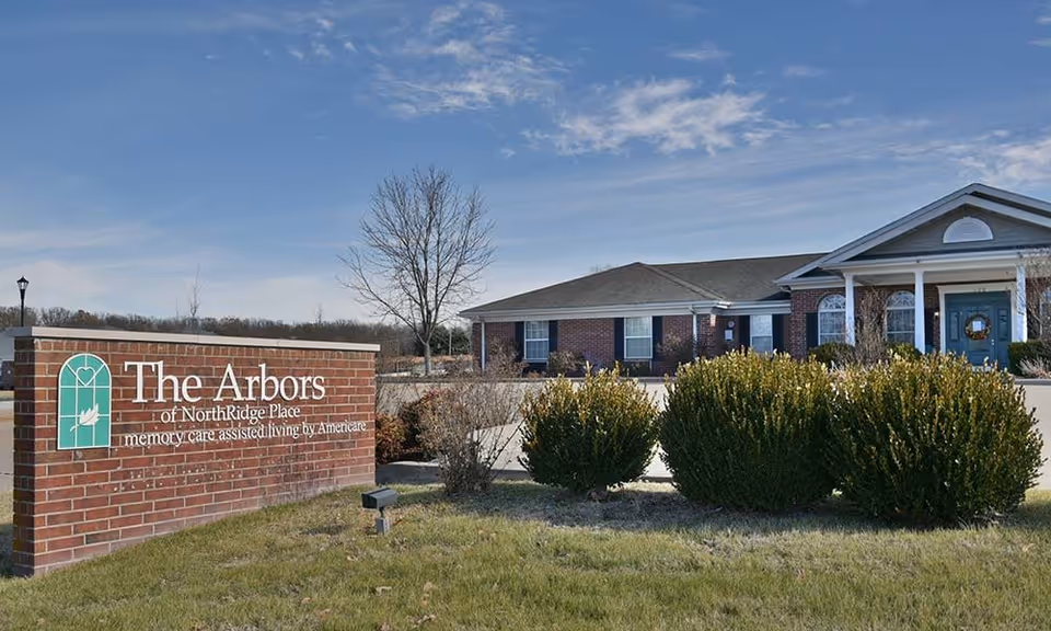 Exterior view of The Arbors of NorthRidge Place, a single-story brick building with a gabled roof and a covered entrance supported by white columns. In front of the building is a landscaped area with bushes and a brick sign that reads 'The Arbors of NorthRidge Place memory care, assisted living by Americare' under a blue sky with scattered clouds.