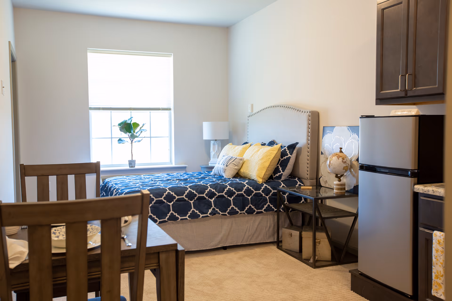 A cozy bedroom with a bed dressed in blue and white patterned bedding and yellow accent pillows. There is a window with blinds letting in natural light, a small potted plant on the windowsill, a bedside table with a lamp, and a glass side table holding a decorative globe and artwork. A small stainless steel refrigerator and dark wood cabinets are visible on the right side. In the foreground, there is a wooden dining table with chairs set with plates and utensils.
