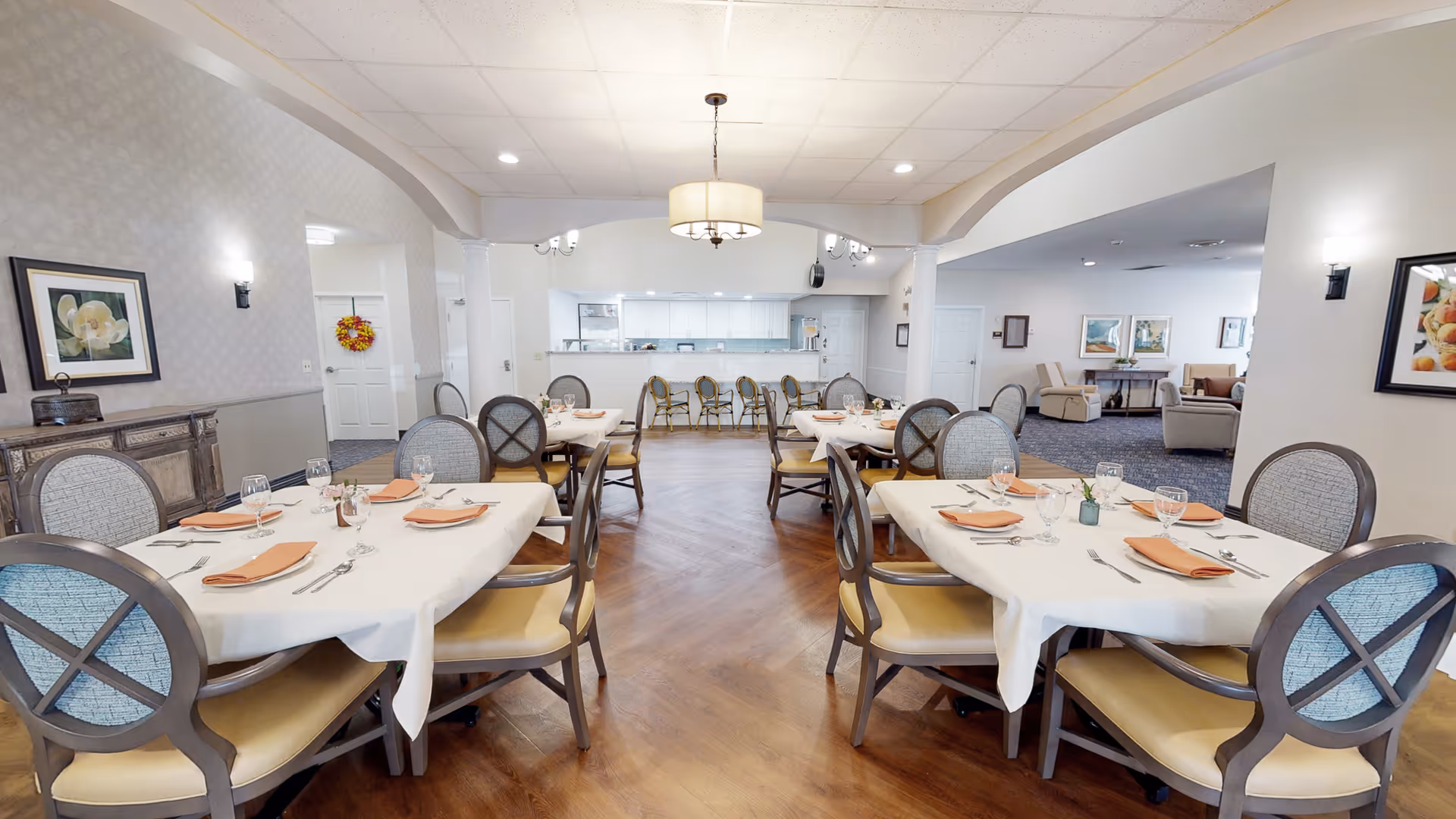 A bright dining room in a senior living facility with several tables covered in white tablecloths, each set with orange napkins, glassware, and silverware. The room features wooden flooring, cushioned chairs with patterned backs, and a large hanging light fixture in the center. In the background, there is a kitchen area with a counter and bar stools, and a lounge area with armchairs and framed artwork on the walls.