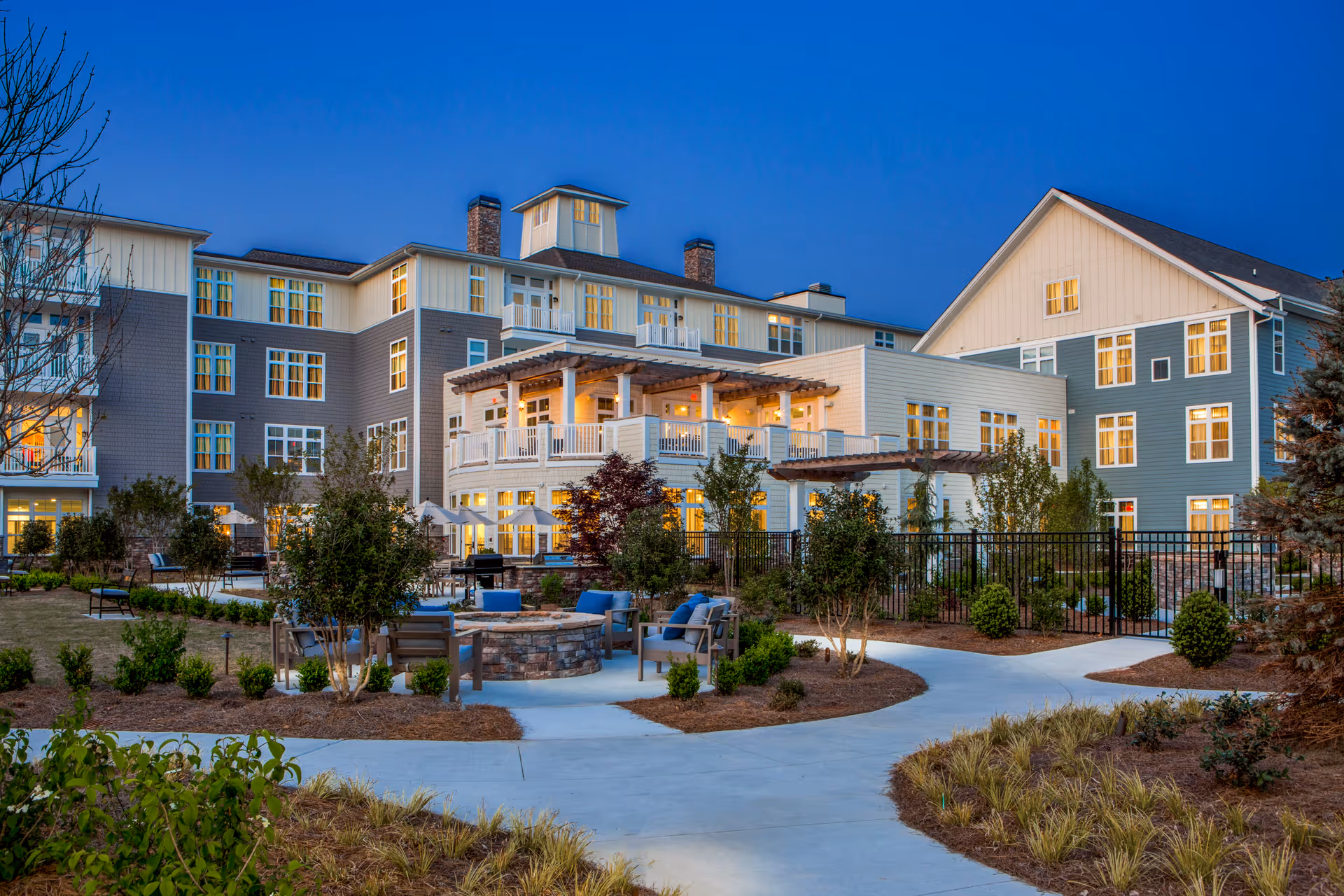 Exterior view of The Arbor at BridgeMill senior living facility at dusk, showing a multi-story building with lit windows, outdoor seating areas with chairs and tables, landscaped gardens, and paved walkways.