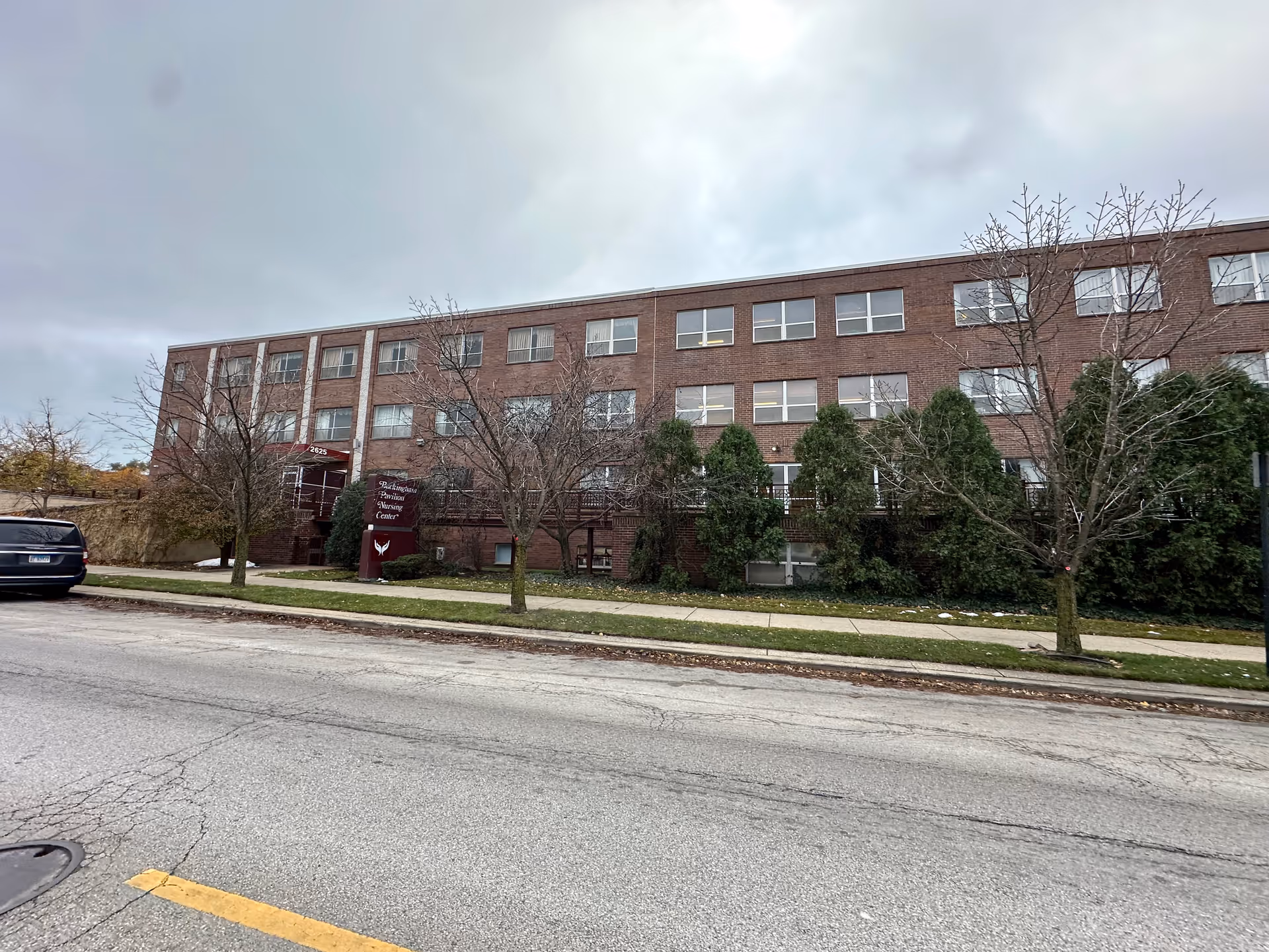 Exterior view of a three-story brick building with multiple windows, a sidewalk, and some leafless trees and bushes in front. A maroon sign near the entrance reads 'Buckingham Pavilion Nursing Center'. A black vehicle is parked on the street in front of the building under a cloudy sky.