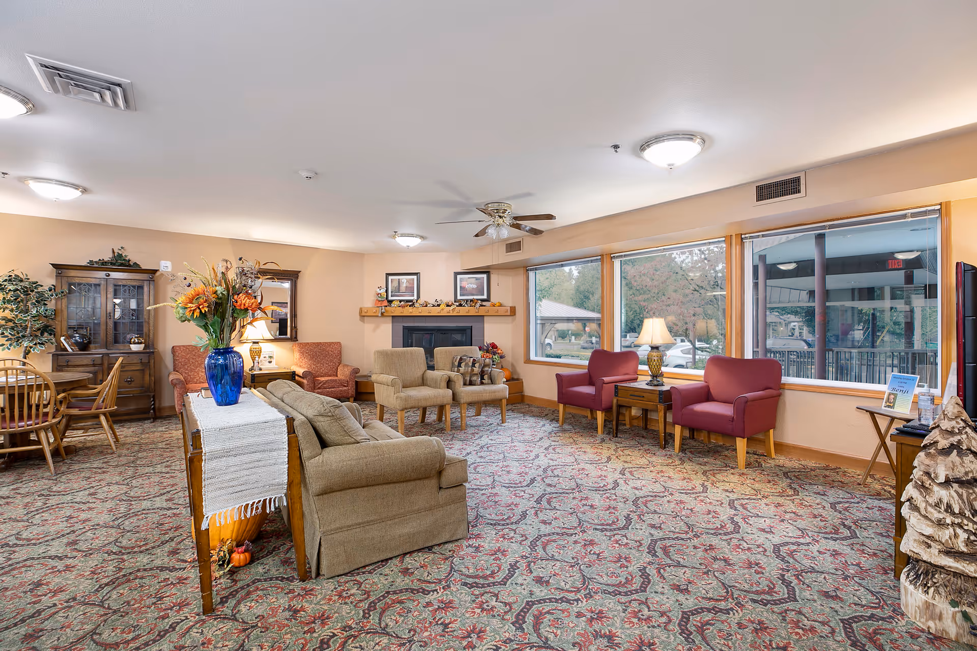 A cozy living room area in a senior living facility with patterned carpet, multiple armchairs in beige and red, a sofa, a wooden table with a blue vase of flowers, a fireplace with decorations above it, large windows showing an outdoor view, and warm lighting.