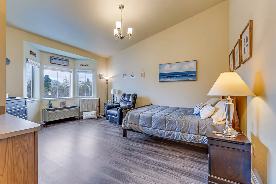 Sunlit studio bedroom with a single bed, armchair, nightstand and a bay window overlooking trees.