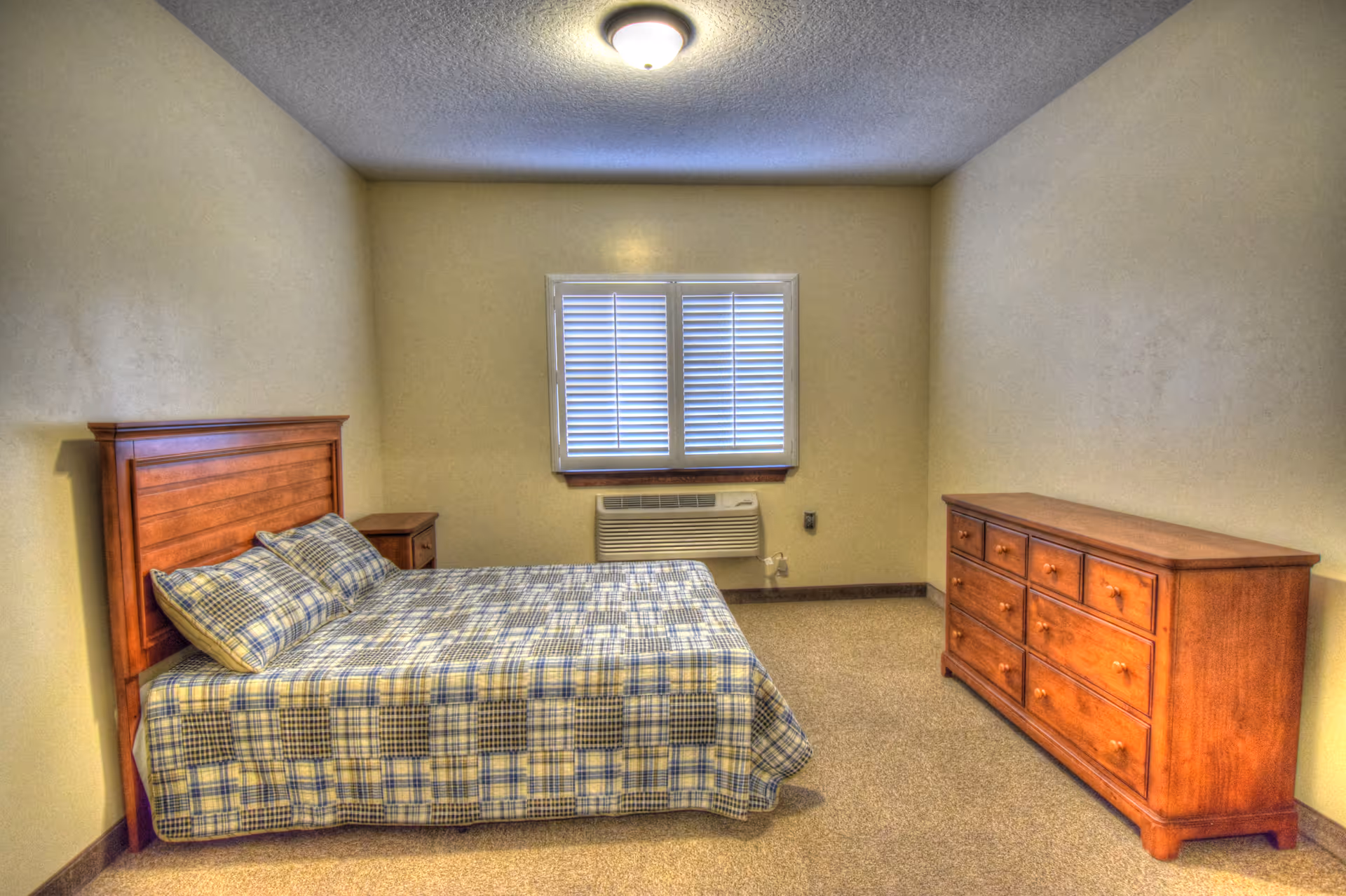A simple bedroom with a wooden bed frame and a plaid bedspread, two matching pillows, a wooden nightstand, a wooden dresser with multiple drawers, a window with closed blinds, and a ceiling light fixture.