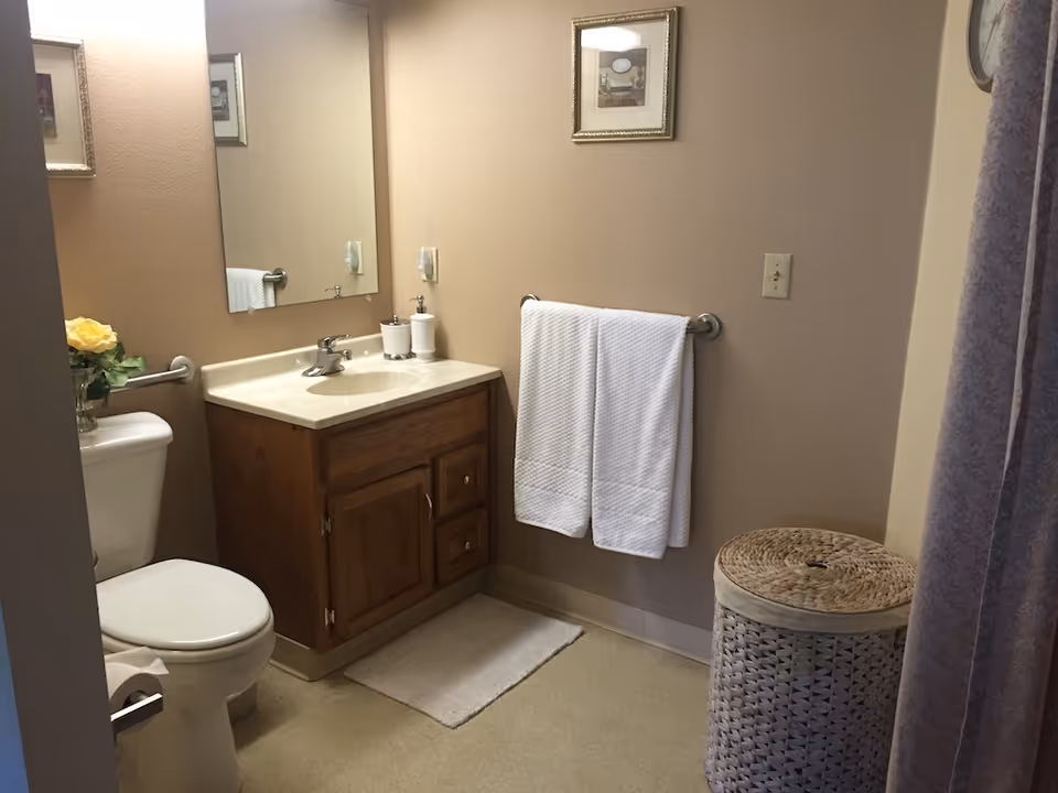 A bathroom with a toilet, wooden vanity with a sink, a large mirror above the sink, a towel rack with two white towels, a wicker laundry basket, and a shower curtain partially visible on the right side. There is a small framed picture on the wall and a vase with yellow flowers on the toilet tank.