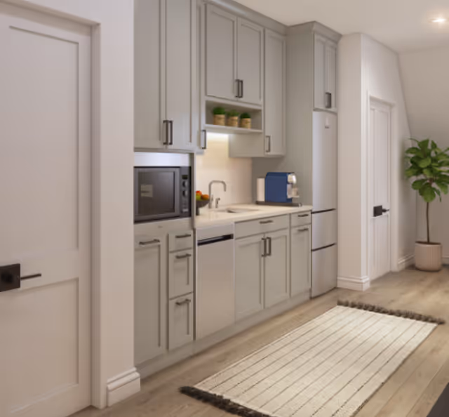 A modern kitchen area with light gray cabinets, a built-in microwave, dishwasher, refrigerator, and a coffee machine on the countertop. There is a small potted plant on the counter and a larger potted plant in the corner near a closed door. A light-colored rug with dark fringes is placed on the wooden floor.