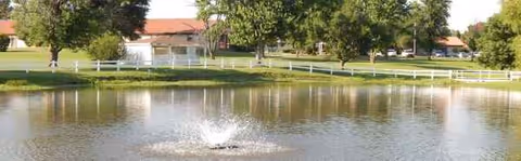 Outdoor pond and fountain at Franciscan Villa, with trees and a distant building.