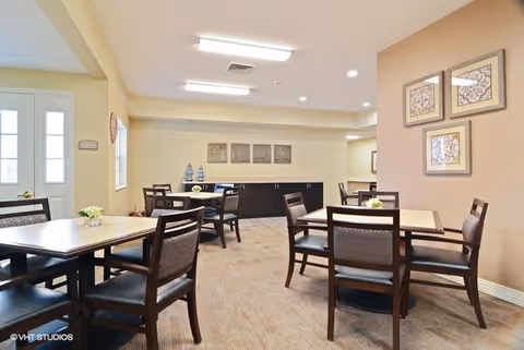 A well-lit dining room with several square wooden tables and cushioned chairs arranged neatly. The walls are painted in neutral tones and decorated with framed artwork. There is a sideboard against the far wall with decorative items on top. A door and window are visible on the left side of the room.