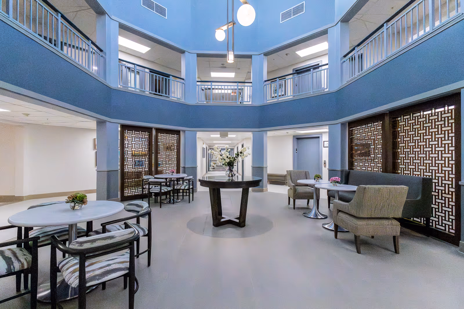 Two-story atrium lounge with round tables, chairs and seating areas around a central table in a senior living facility.