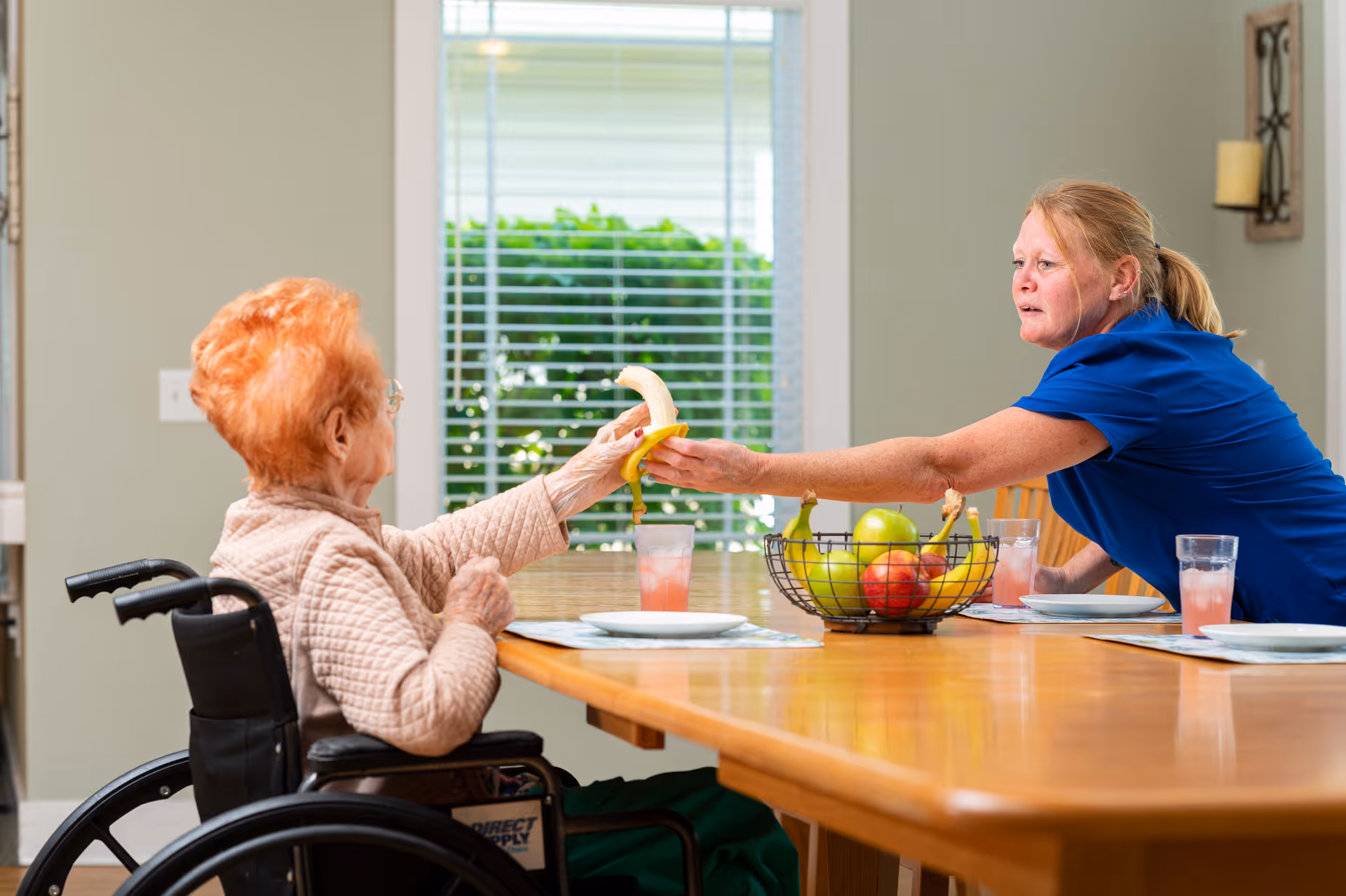 An elderly woman in a wheelchair is sitting at a dining table inside a room, reaching out to receive a peeled banana from a caregiver wearing a blue shirt. The table has a fruit basket with apples and bananas, placemats, plates, and glasses with pink beverages. A window with blinds and greenery outside is visible in the background.