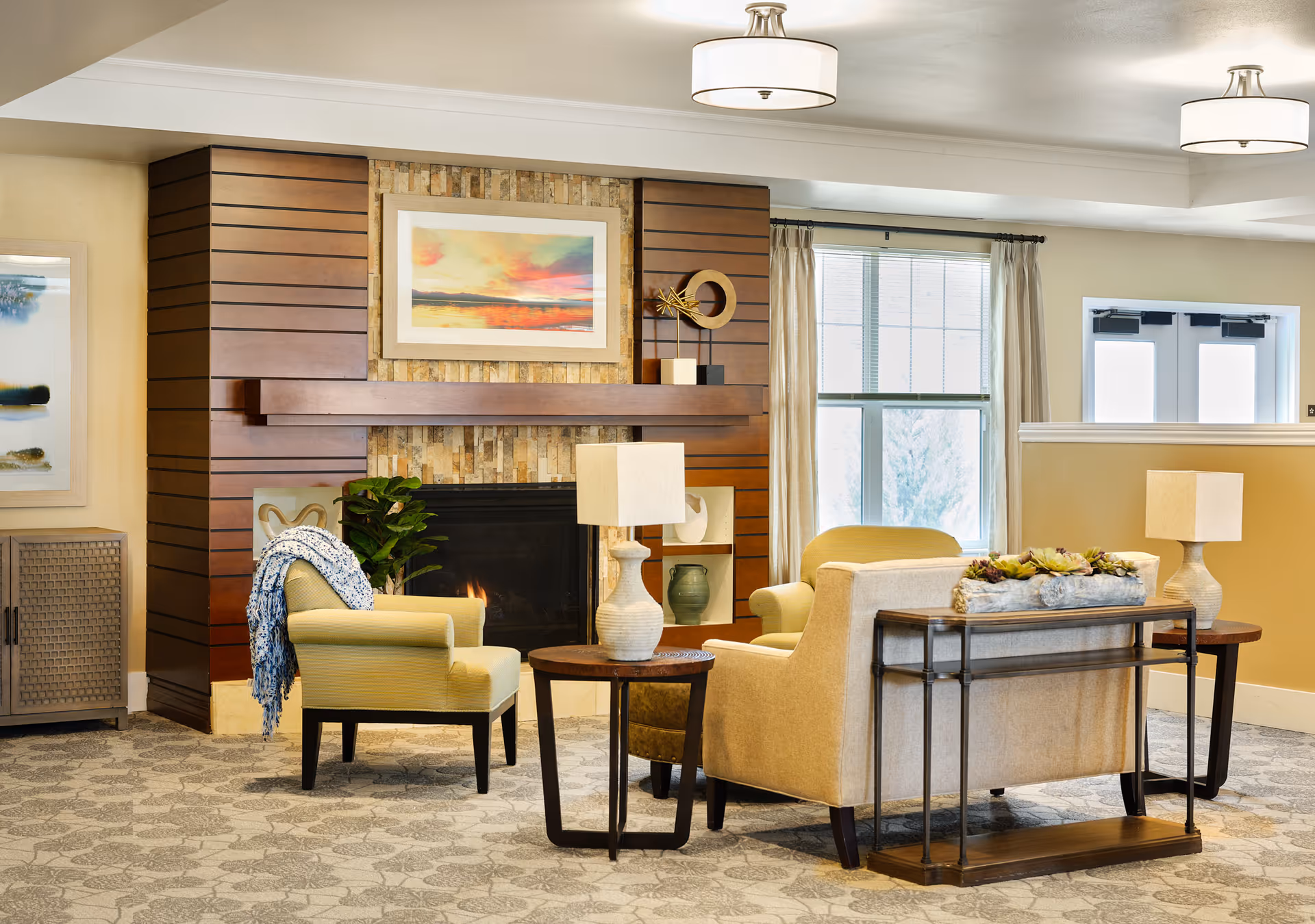 A cozy living room area with a fireplace surrounded by wooden paneling and stone. The room features comfortable armchairs, a sofa, side tables with lamps, decorative plants, and framed artwork above the fireplace. Large windows with curtains allow natural light to fill the space.