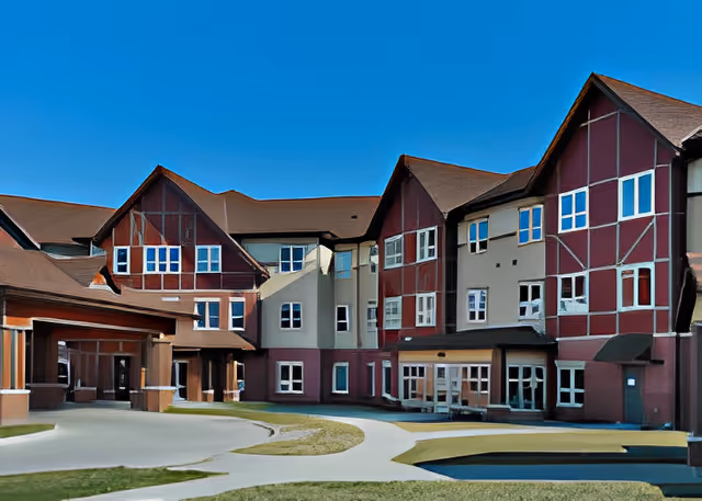 Exterior view of a large multi-story senior living facility building with red and beige walls, multiple windows, and a clear blue sky above. There is a paved pathway and some grass in front of the building.