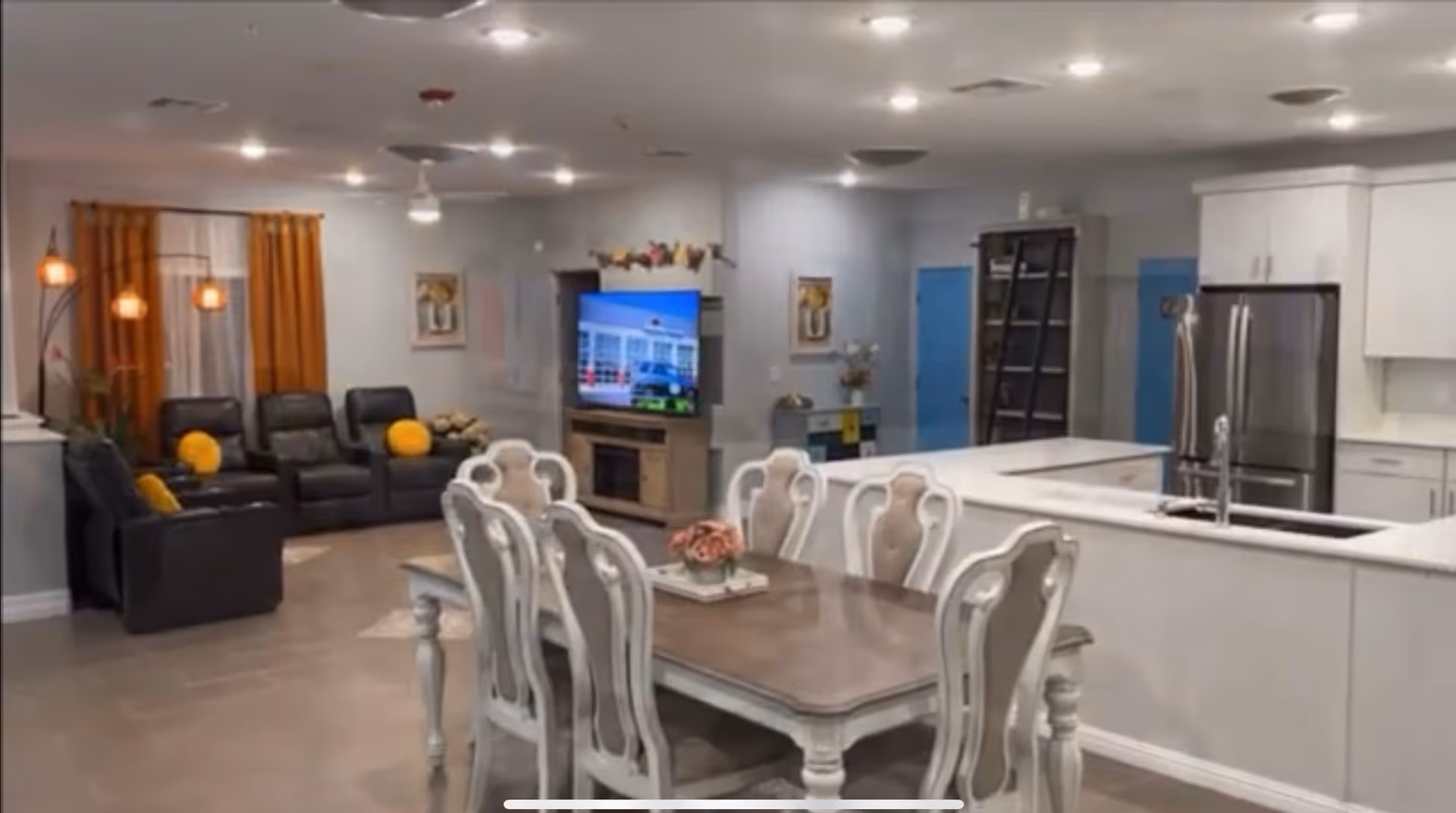 Interior view of a residential assisted living facility showing a combined living and dining area. The foreground features a wooden dining table with six white chairs. In the background, there is a living room area with black leather recliners and yellow cushions, a TV mounted on a stand, and orange curtains. To the right, a modern kitchen with white cabinets, a stainless steel refrigerator, and a sink is visible.