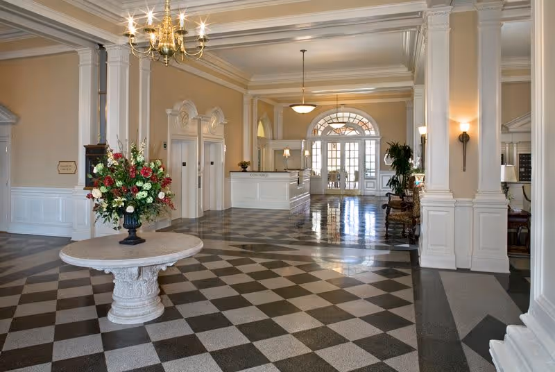 Spacious and elegant lobby area with checkered floor tiles, a round table with a large floral arrangement in the center, white paneled walls, two elevators, a reception desk, and large windows with glass doors letting in natural light.