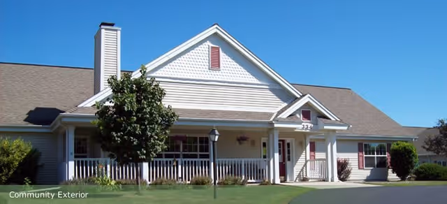 Exterior view of a single-story assisted living facility building with a gabled roof, white siding, a front porch with railings, a tree, and a lamp post on a well-maintained lawn under a clear blue sky.