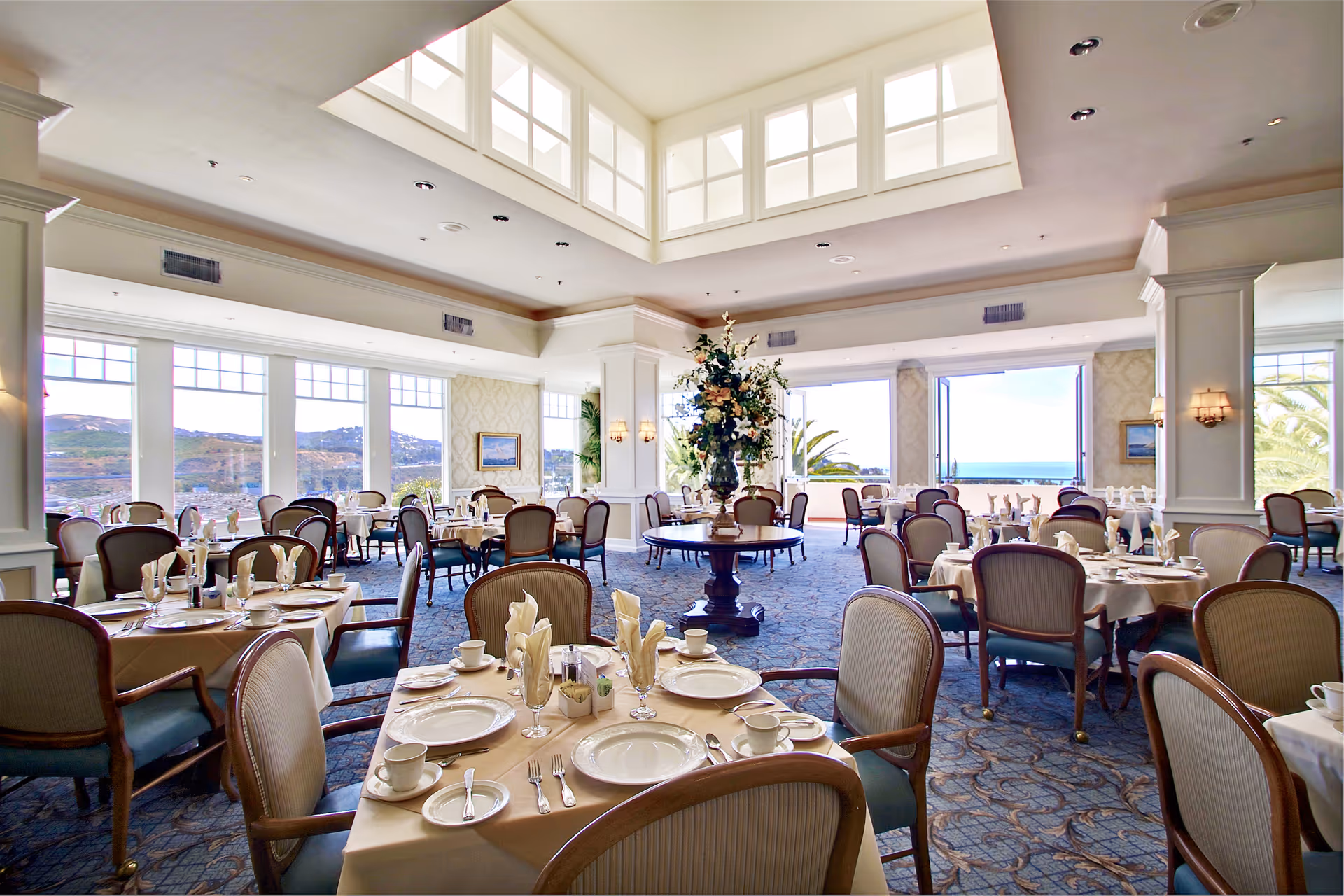 A bright and elegant dining room with multiple tables set with plates, cups, and silverware. The room features large windows offering scenic views of hills and the ocean, with a central round table adorned with a large floral arrangement. The ceiling has a skylight, and the room is decorated with light-colored walls and carpeted floors.