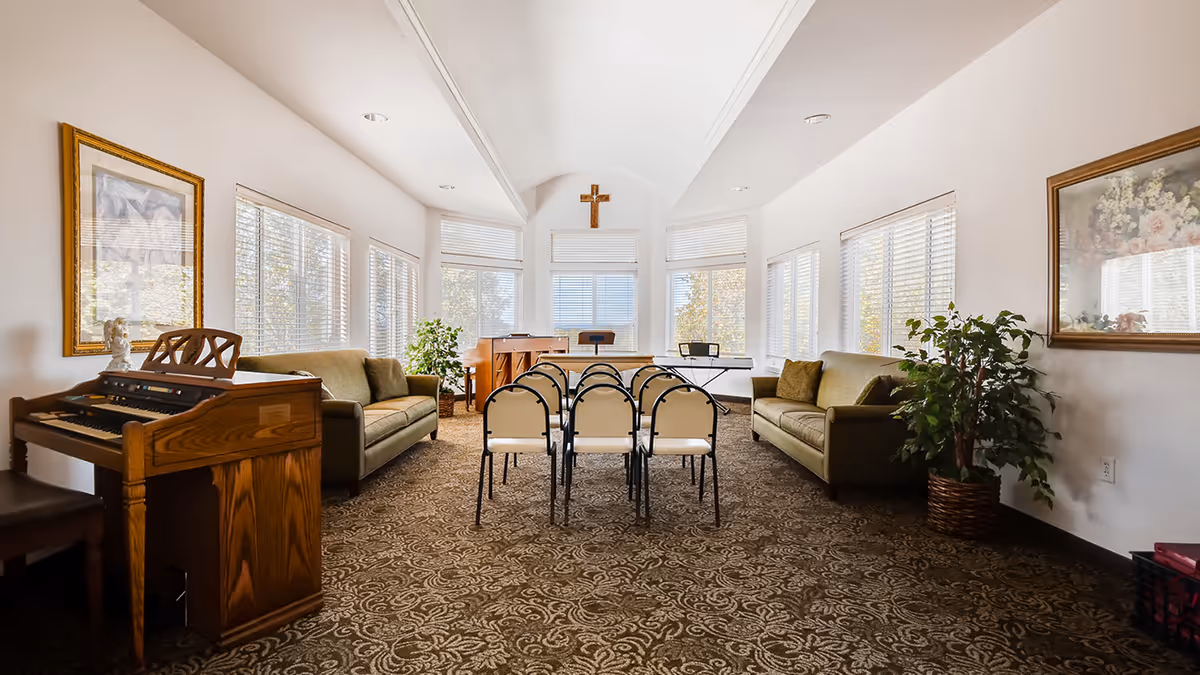 Sunlit common room with rows of folding chairs facing a small altar with a cross, flanked by sofas, an organ, and potted plants.