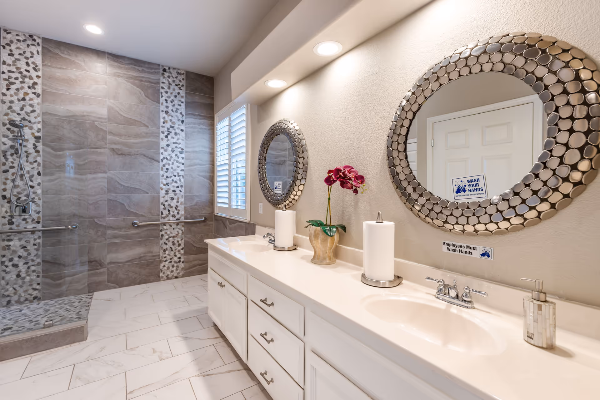 A modern bathroom with a double sink vanity featuring white countertops and cabinets, two round decorative mirrors above the sinks, a paper towel holder, a soap dispenser, and a potted orchid. The shower area has large gray tiles with pebble mosaic accents and a glass door. The floor is tiled with white marble-patterned tiles. There are grab bars installed near the shower for accessibility.