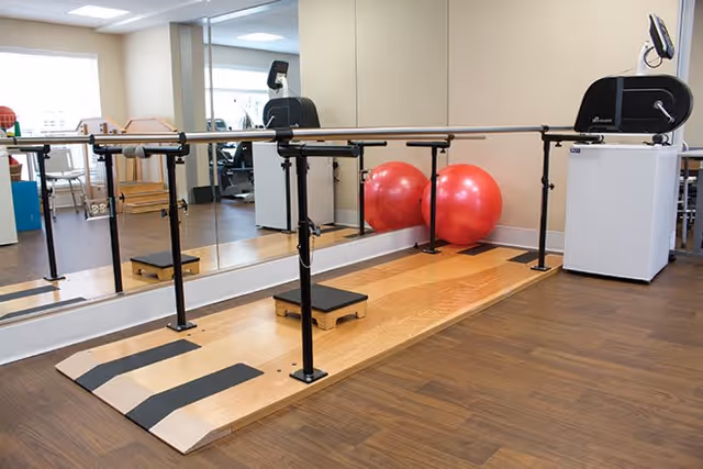 Physical therapy or exercise room with parallel bars for walking support, a small step platform, two large red exercise balls, and a mirrored wall reflecting the equipment and part of the room.